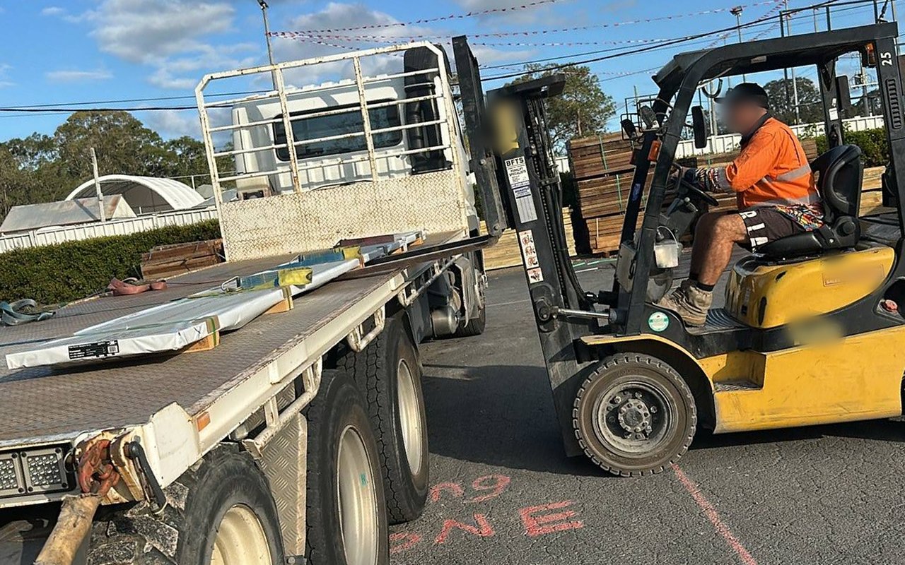 A Dingo tray truck being loaded with timber by a forklift in a supplier's yard